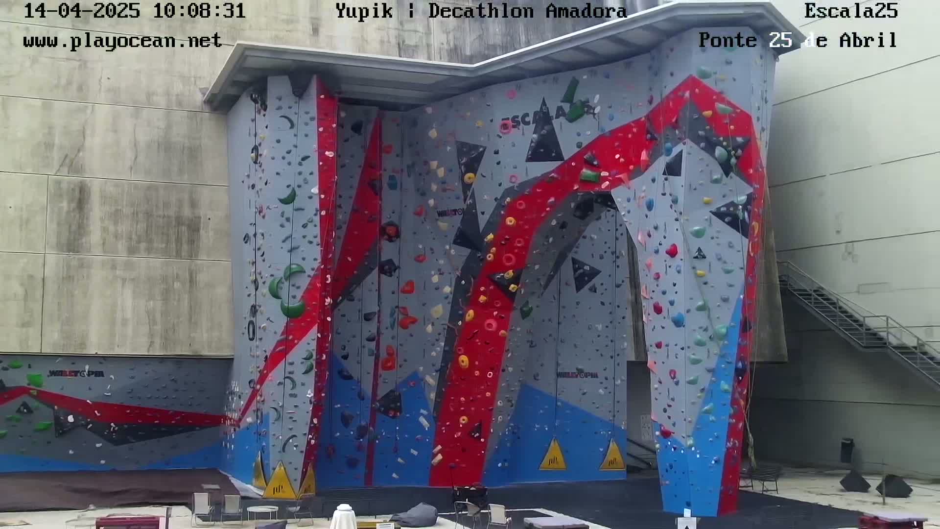 An outdoor climbing wall with various colored holds is situated against a light-grey building on a sunny day.