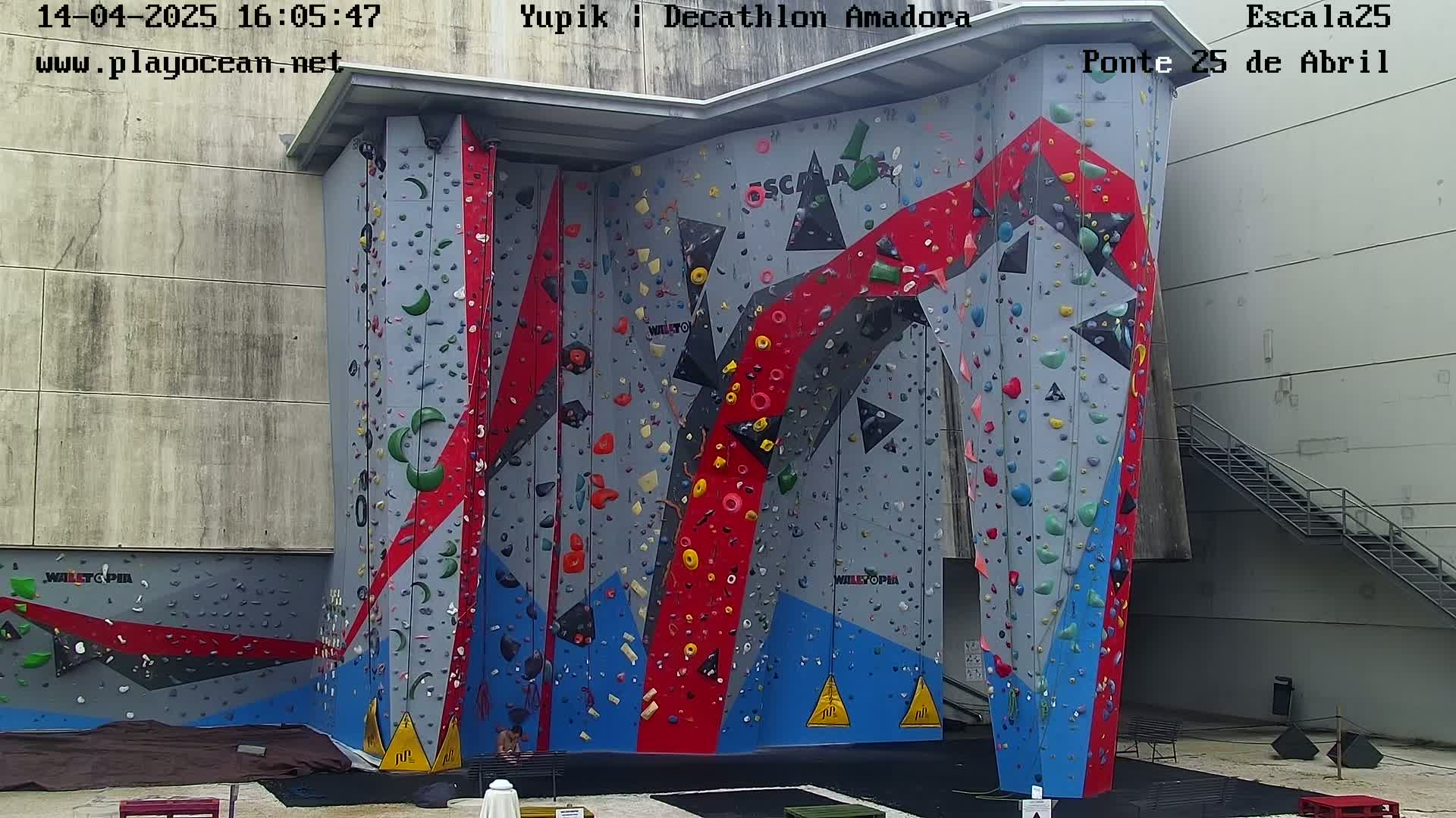 A large, multi-colored climbing wall with various handholds and footholds stands outdoors against a light gray building.