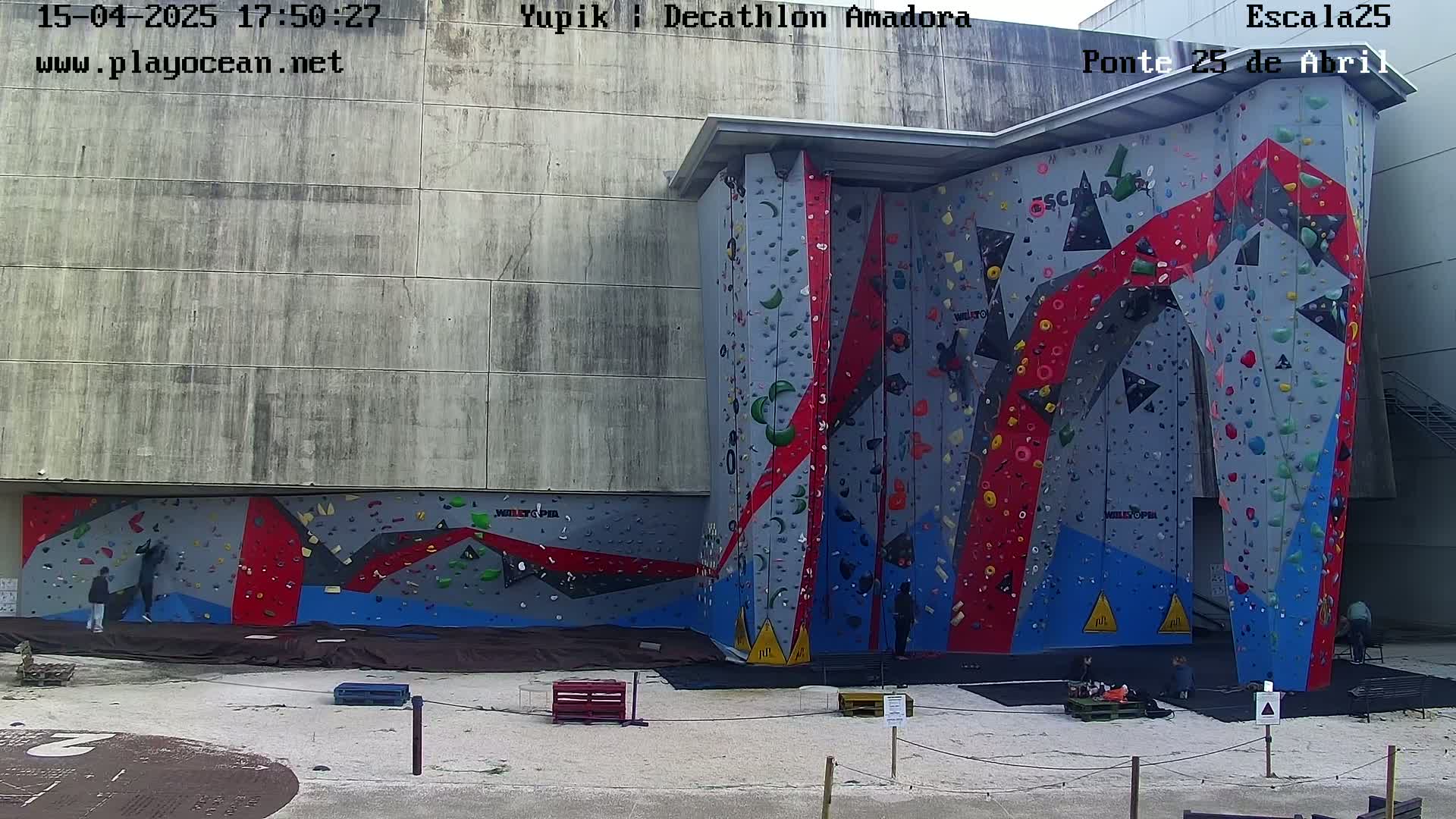 An outdoor climbing wall with various colored holds is situated against a large gray building, under a bright, sunny sky.