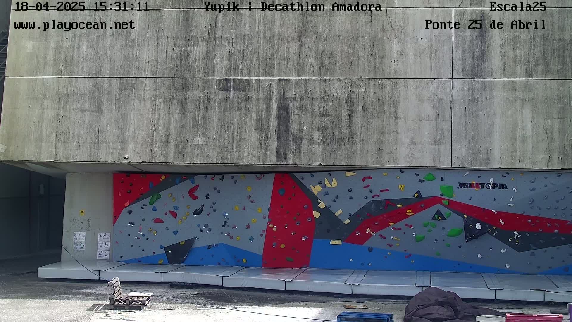 A large outdoor climbing wall with various colored holds is situated beneath a concrete overhang on a sunny day.