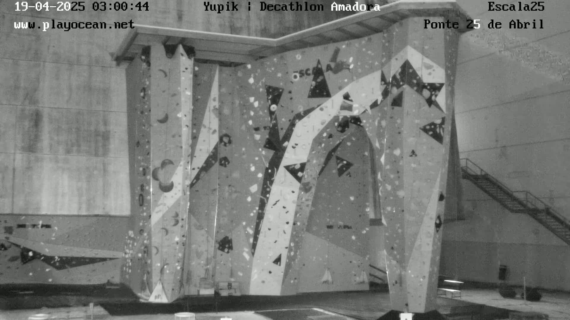A grayscale indoor image shows a large climbing wall with various holds and patterns against a plain wall.