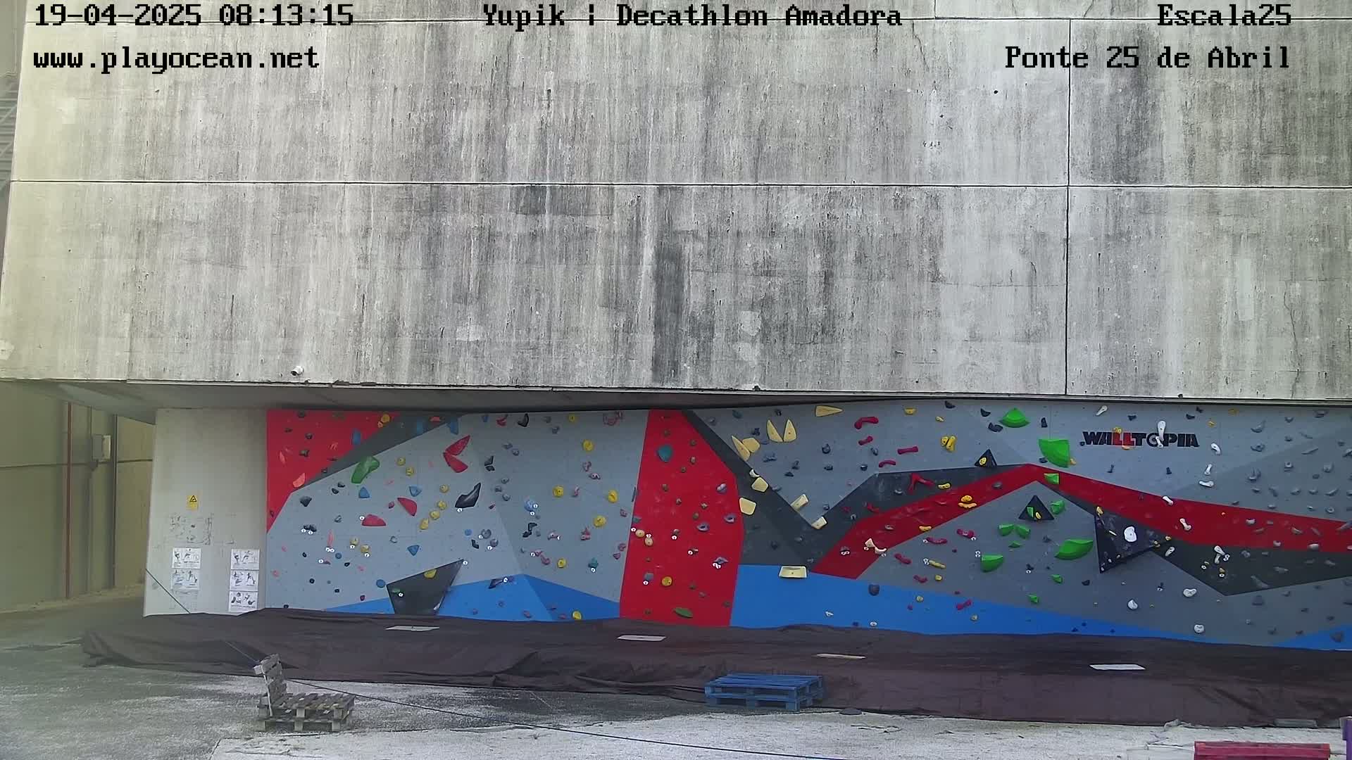 A large outdoor climbing wall with grey, red, and blue sections is situated beneath a concrete overhang.