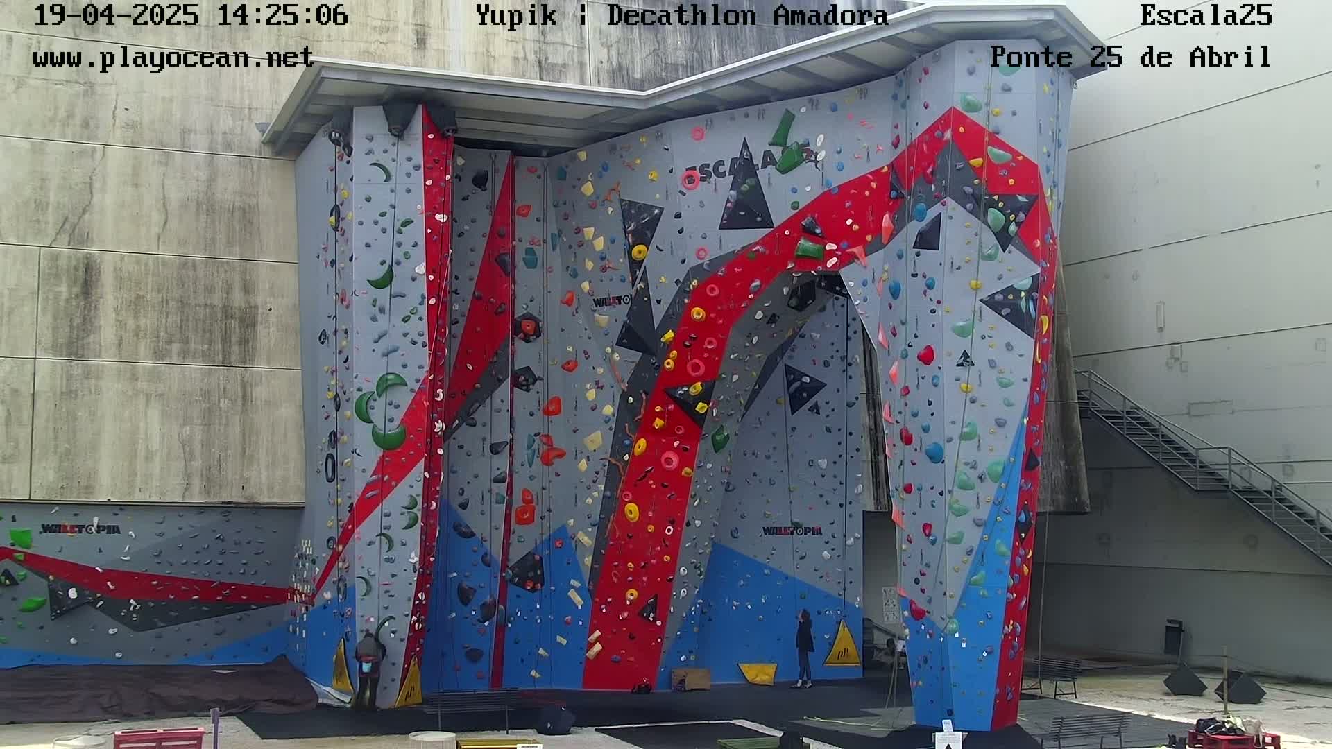 A large, colorful indoor climbing wall with various holds and routes stands against a light gray building on a sunny day.