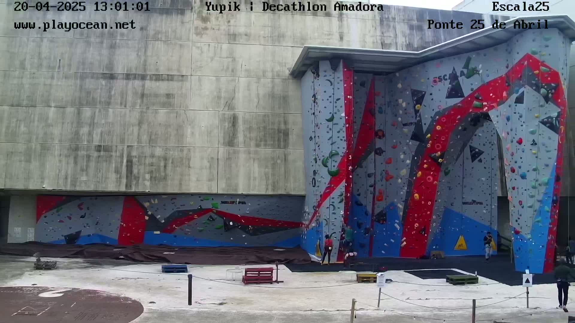 A large outdoor climbing wall with various colored holds is adjacent to a concrete building, with a few people climbing and others standing nearby.
