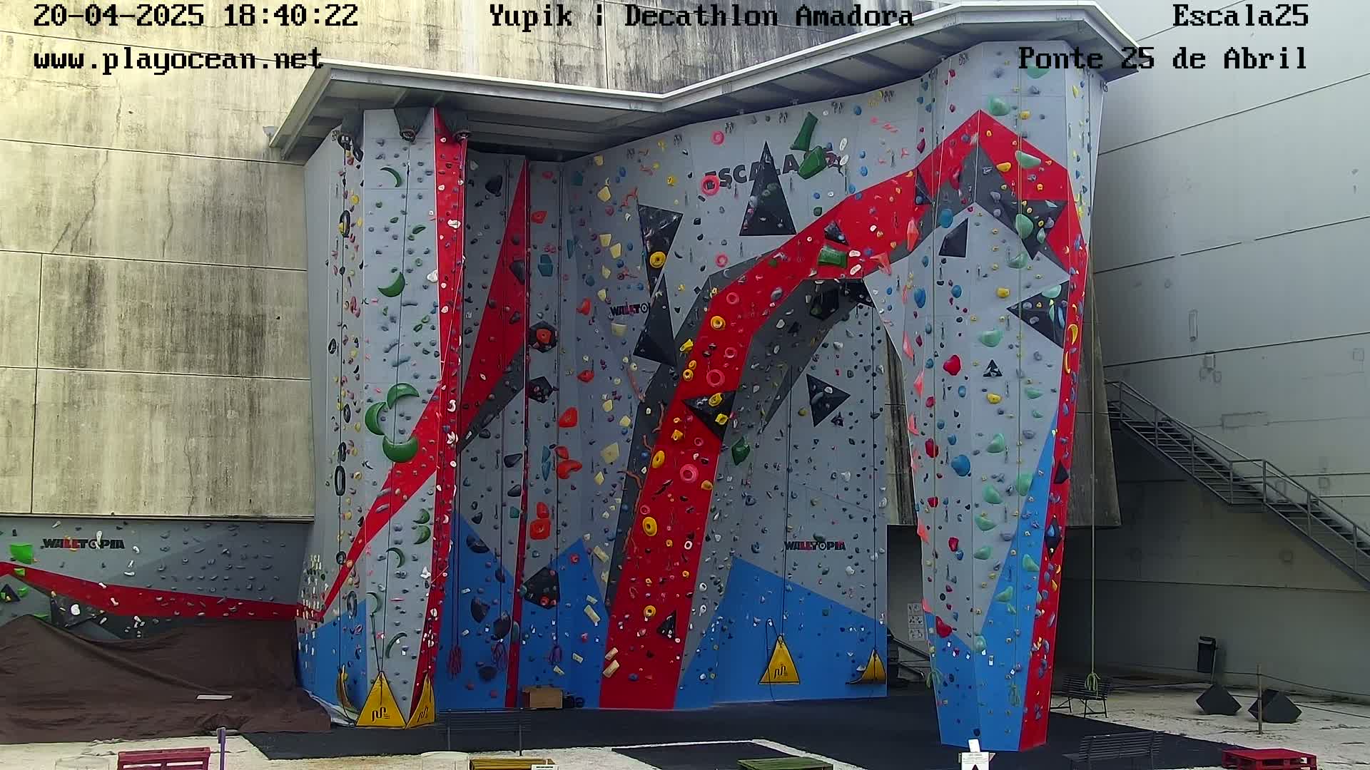 A large, multi-colored indoor climbing wall with various holds is situated against a light gray building.