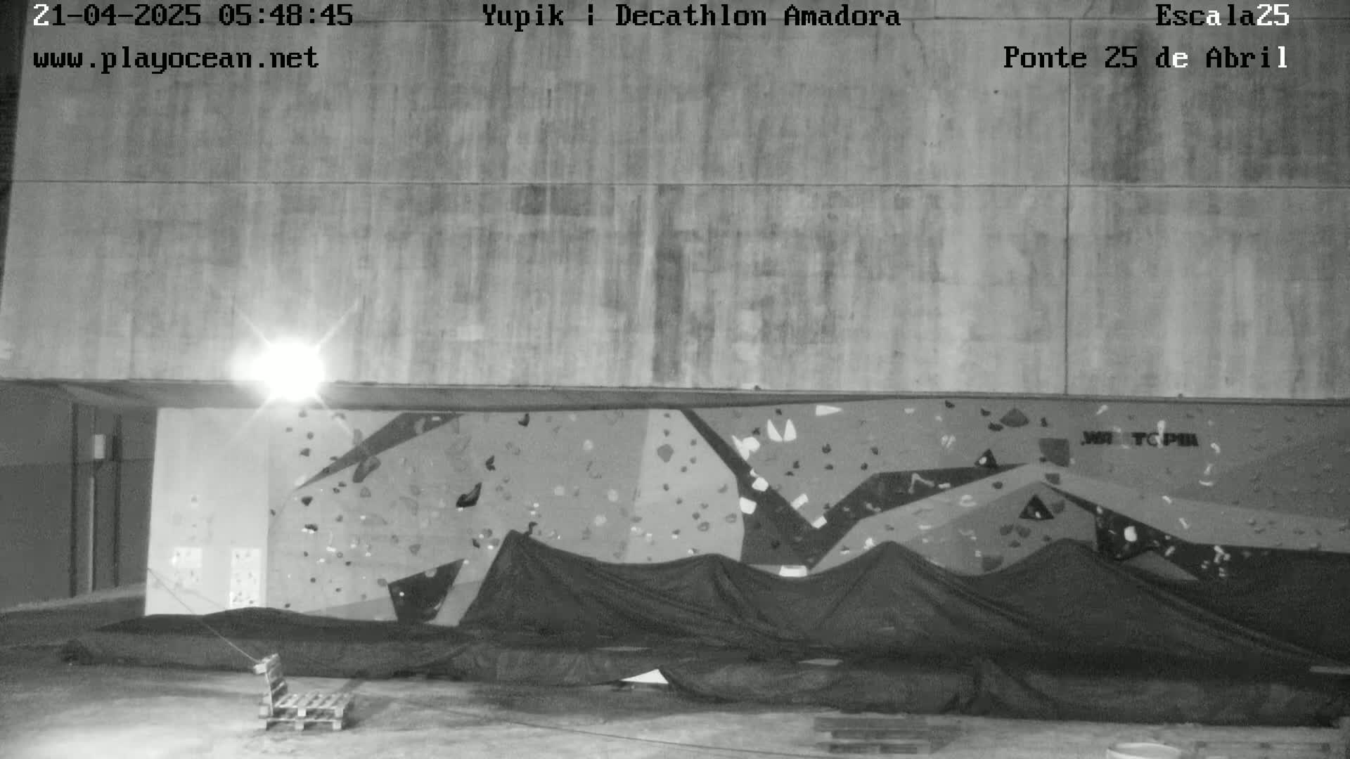 A nighttime black and white image shows an indoor climbing wall covered with dark fabric, under a concrete ceiling with a single light source.
