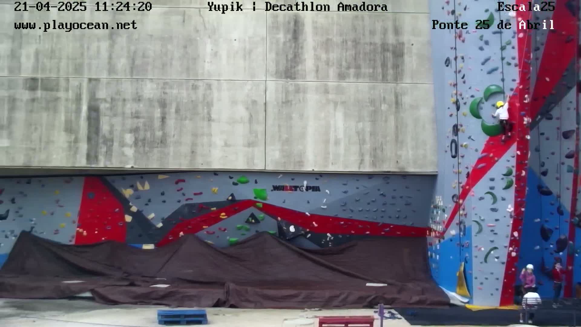 A person climbs a large, colorful outdoor climbing wall, while others watch from below on a partly cloudy day.