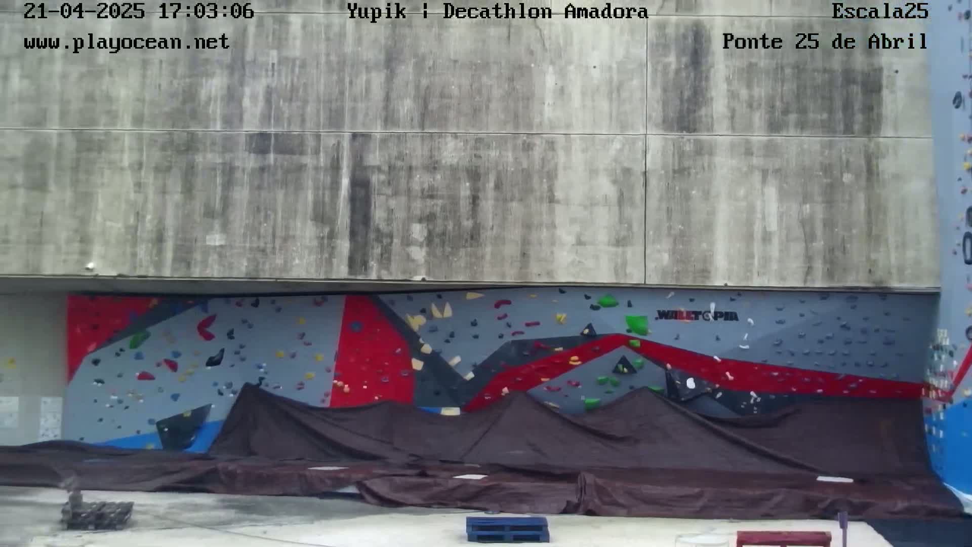 A large, partially covered outdoor climbing wall with red, gray, and blue sections is visible, situated against a concrete wall.