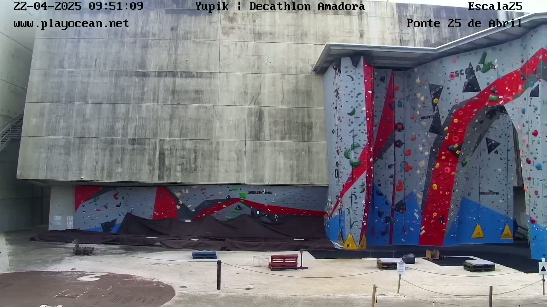 A large outdoor climbing wall with red, blue, and gray holds is situated against a concrete building.