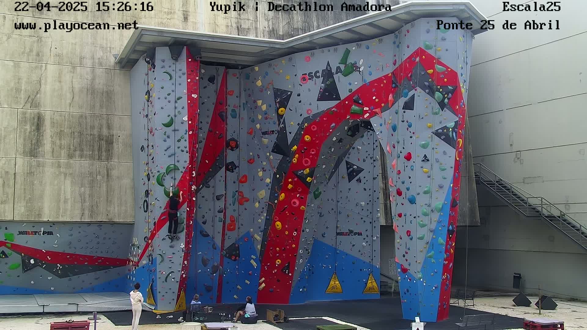 An outdoor climbing wall with various colored holds is positioned against a light gray building, with two people standing near it.