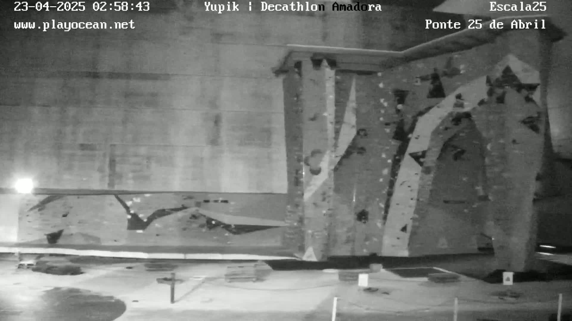 A nighttime, grayscale image shows an outdoor climbing wall with various holds affixed to its surface.