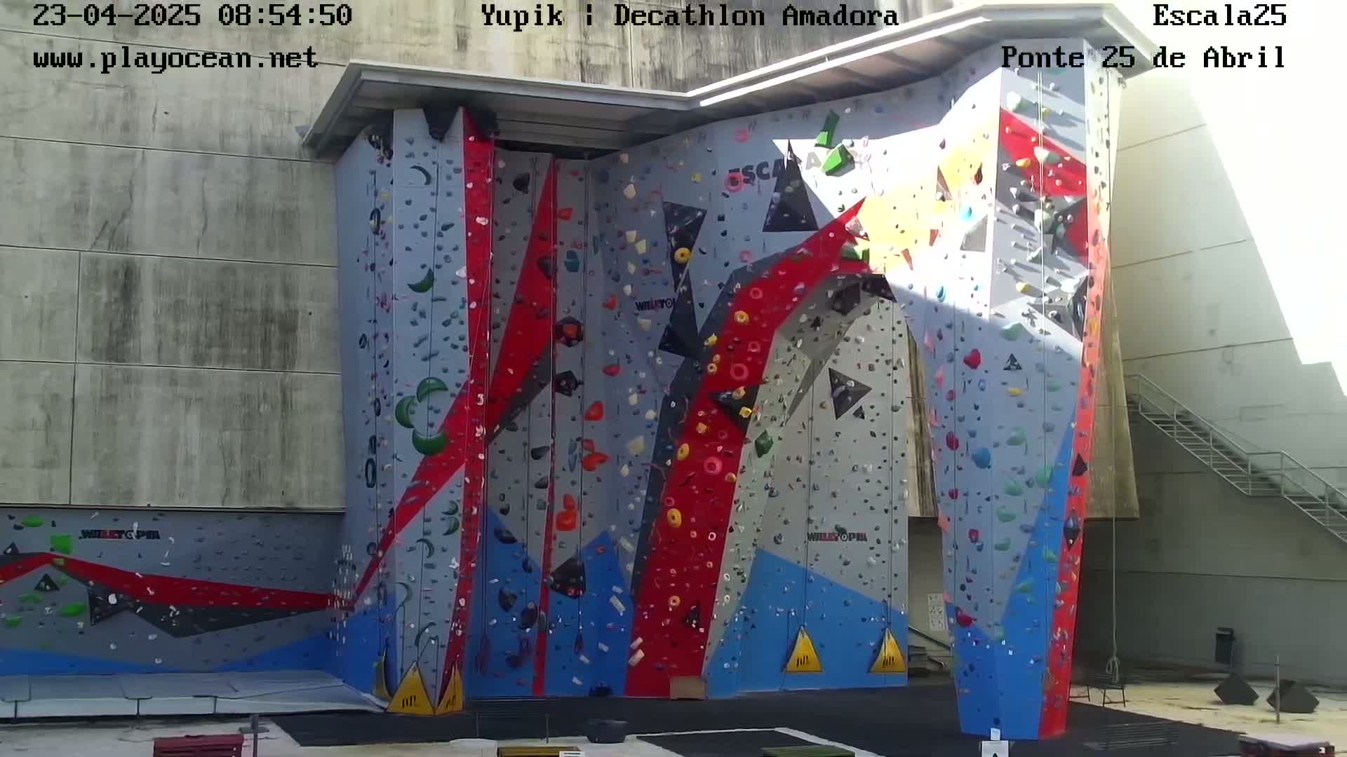 A large, colorful indoor climbing wall stands against a light gray building on a sunny day.