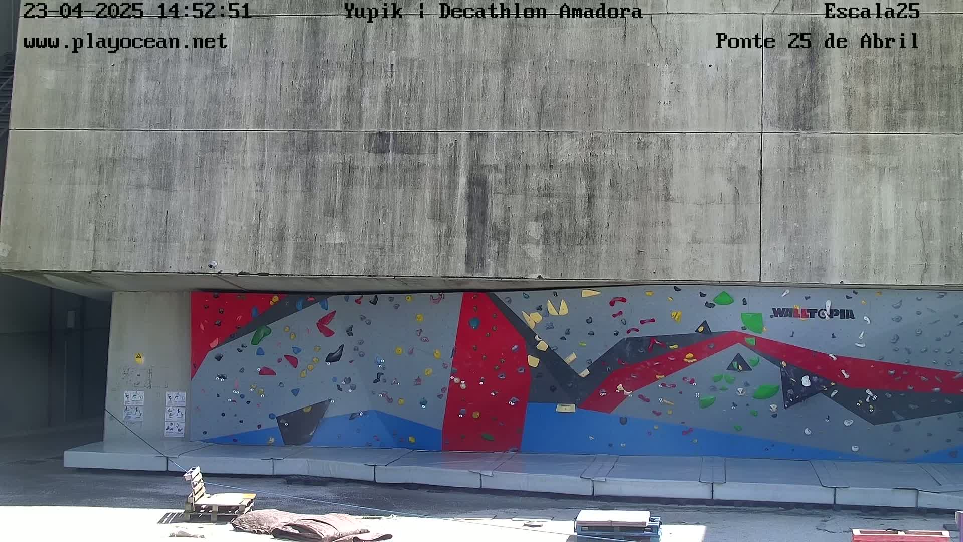 A large, multicolored climbing wall is situated beneath a concrete overhang outdoors in sunny weather.