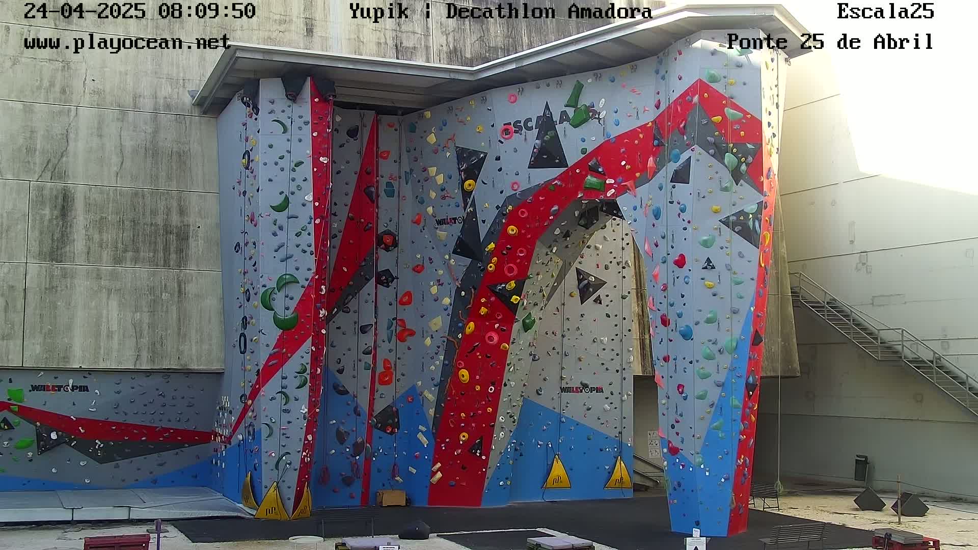 A large, colorful indoor climbing wall with various holds stands against a light-grey building on a sunny day.
