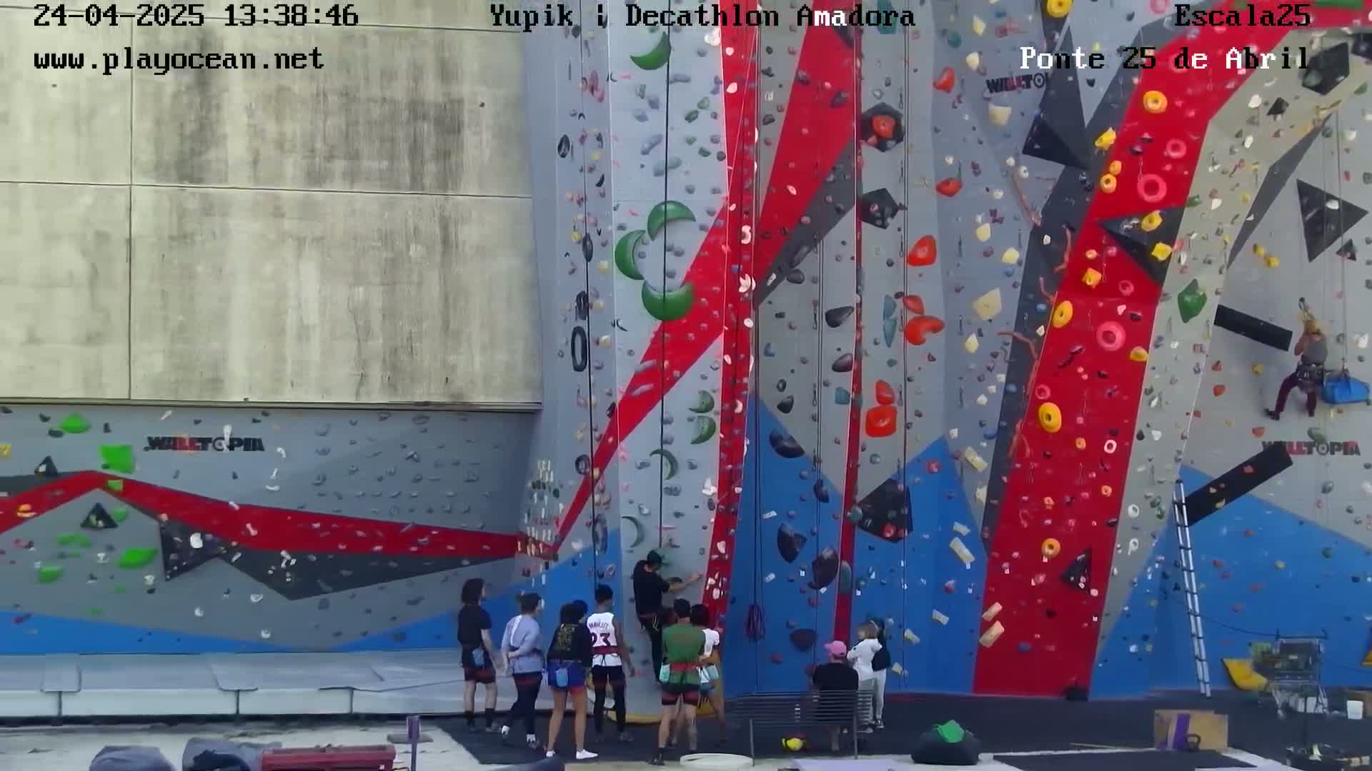 A large, colorful outdoor climbing wall with several people climbing and others watching on a cloudy day.
