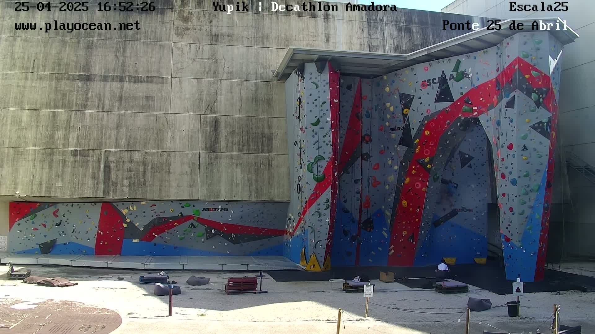A large, colorful climbing wall is situated outside against a concrete building on a sunny day.
