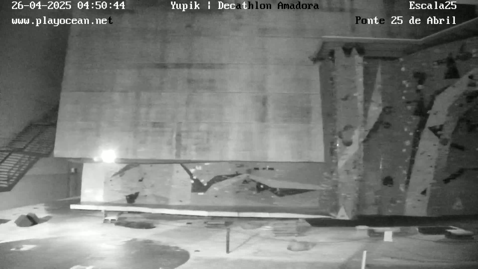A low-light indoor view of a climbing wall with some climbing holds and a metal staircase visible in the background.