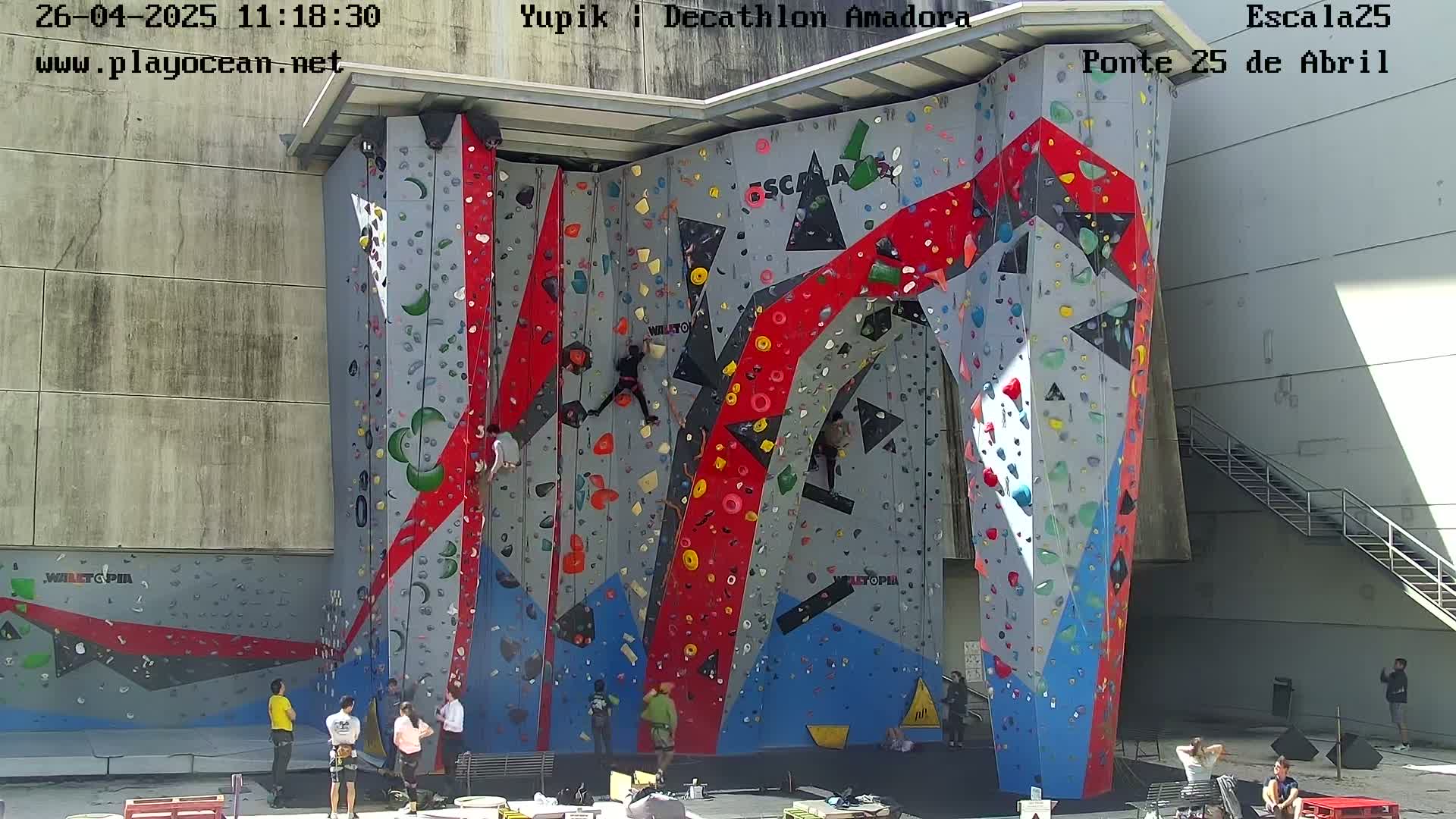 Several people are rock climbing on a large, colorful outdoor climbing wall on a sunny day.