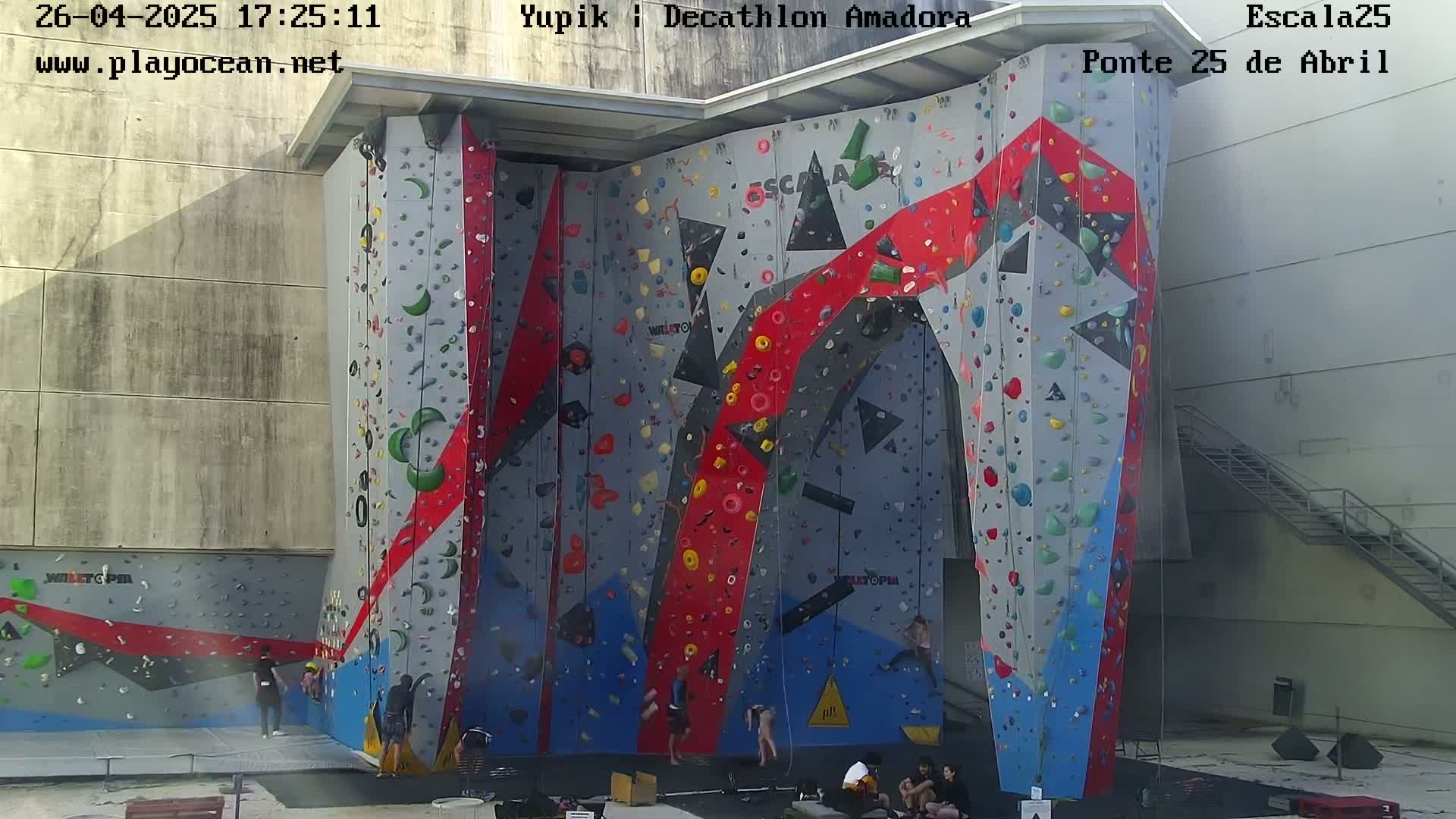 A large, multi-colored climbing wall stands outdoors in bright sunlight, with several people climbing and others sitting at its base.