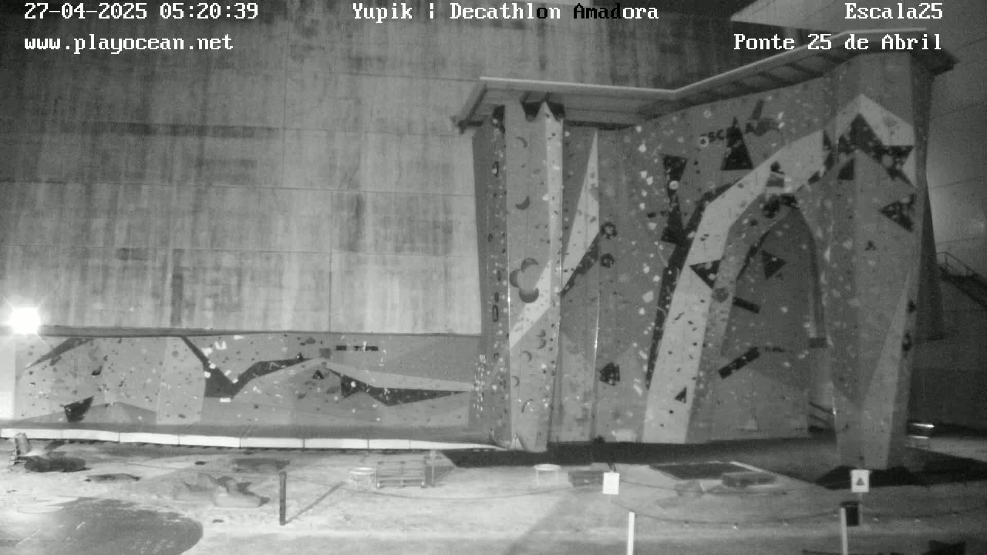 A nighttime black-and-white image shows an indoor climbing wall with various holds and patterns.