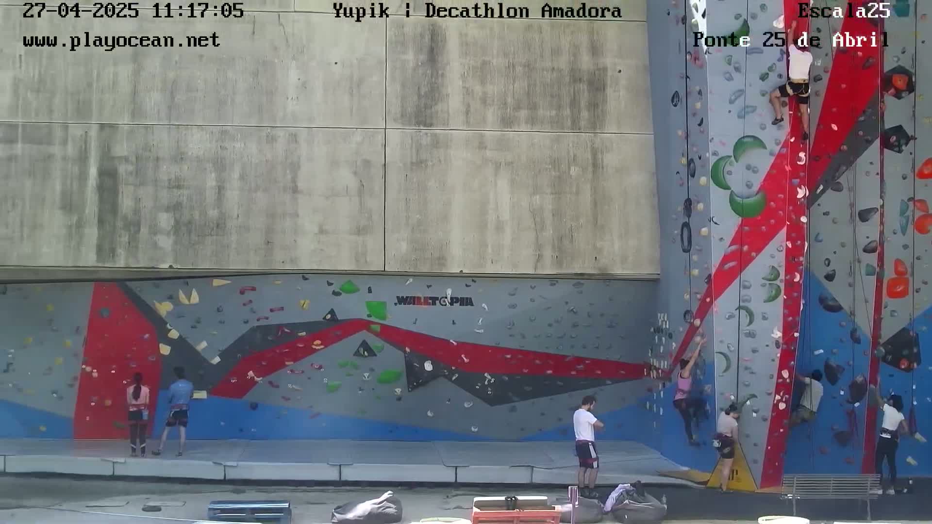 Several people are rock climbing on a large, colorful outdoor climbing wall on a sunny day.