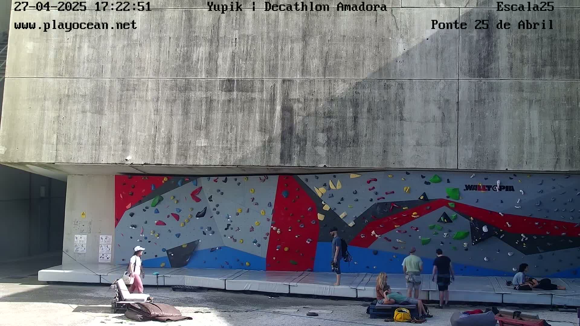 A large, colorful climbing wall is situated outdoors beneath a concrete overhang, with several people standing and sitting near it on a sunny day.