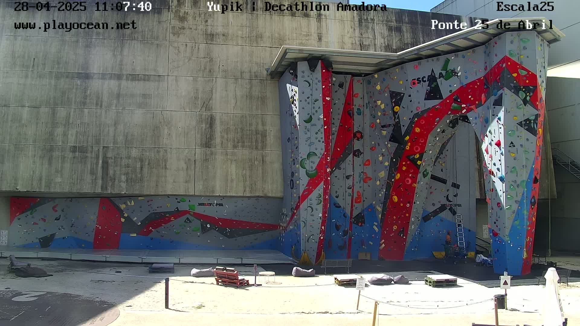 A large, colorful outdoor climbing wall is situated against a light gray building on a sunny day.