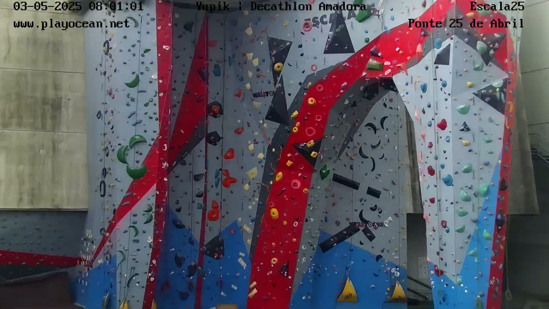 A large indoor climbing wall with various colored holds on a gray, red, and blue surface.
