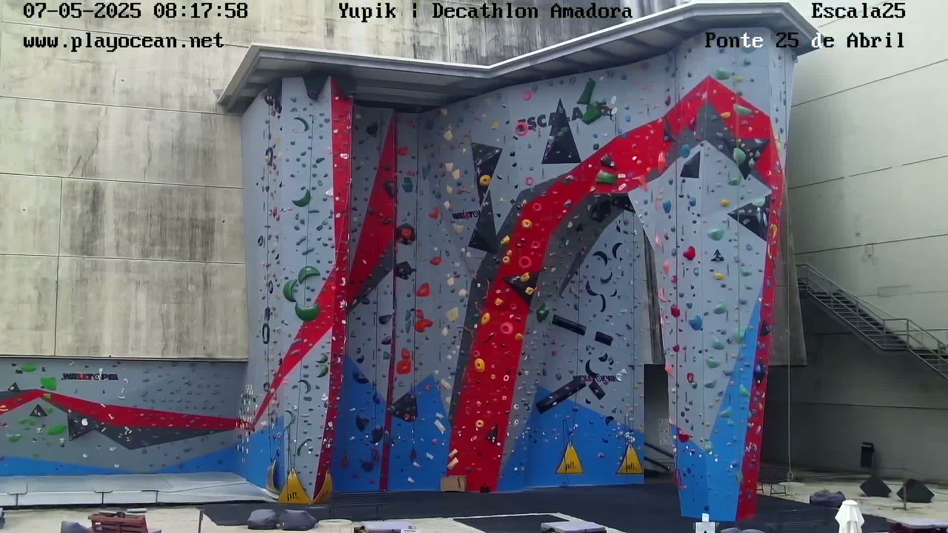 A large, colorful indoor climbing wall with various holds is situated against a light-grey building.