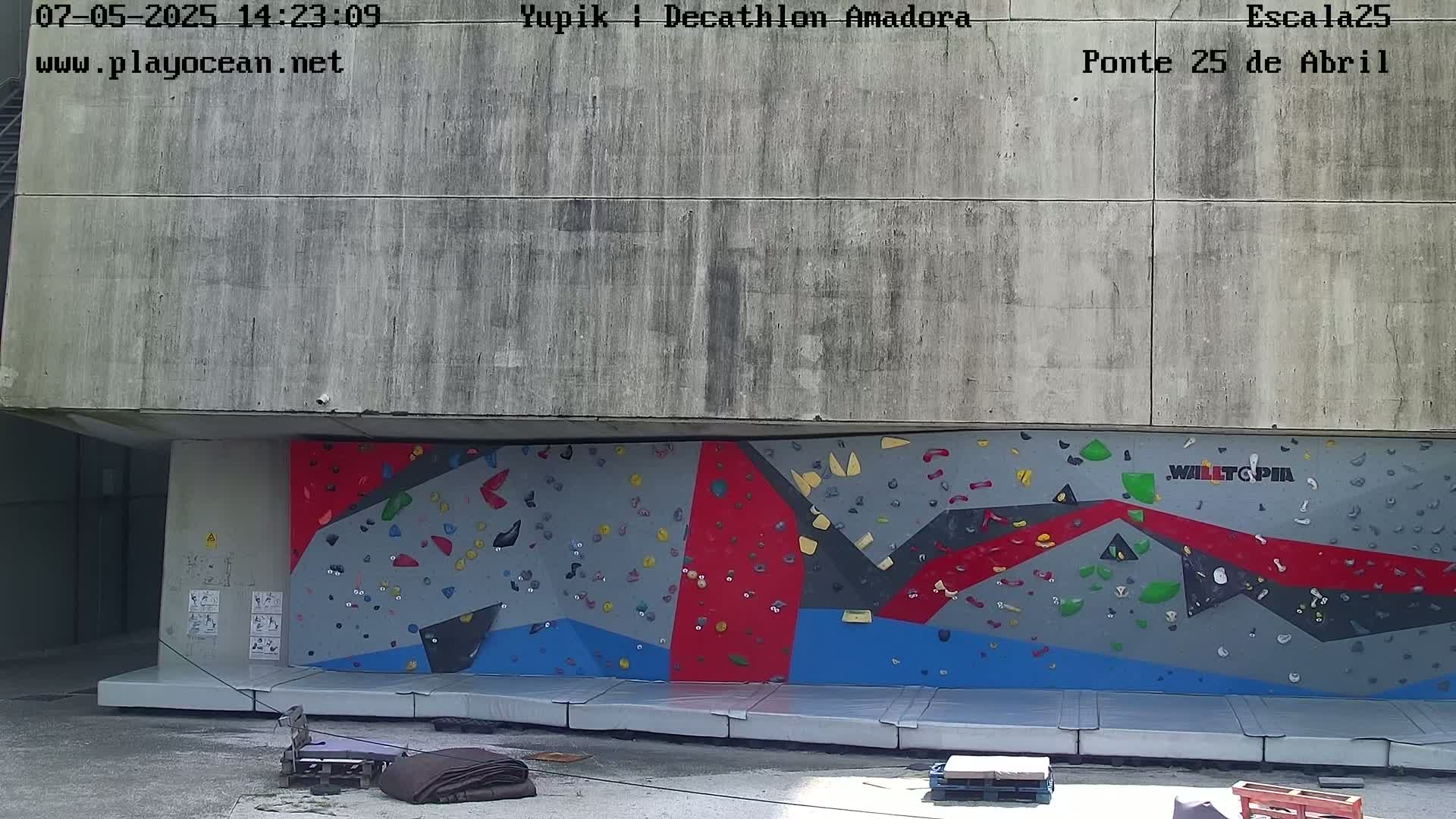 A large outdoor climbing wall with various colored holds is situated against a concrete building under sunny conditions.