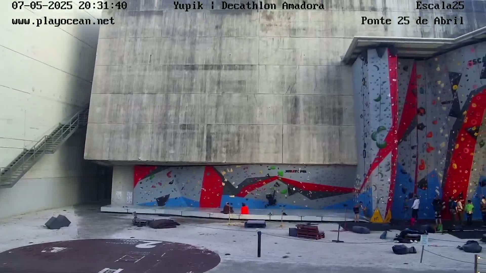 A large outdoor climbing wall with several people climbing and others watching, situated next to a concrete building with a staircase.
