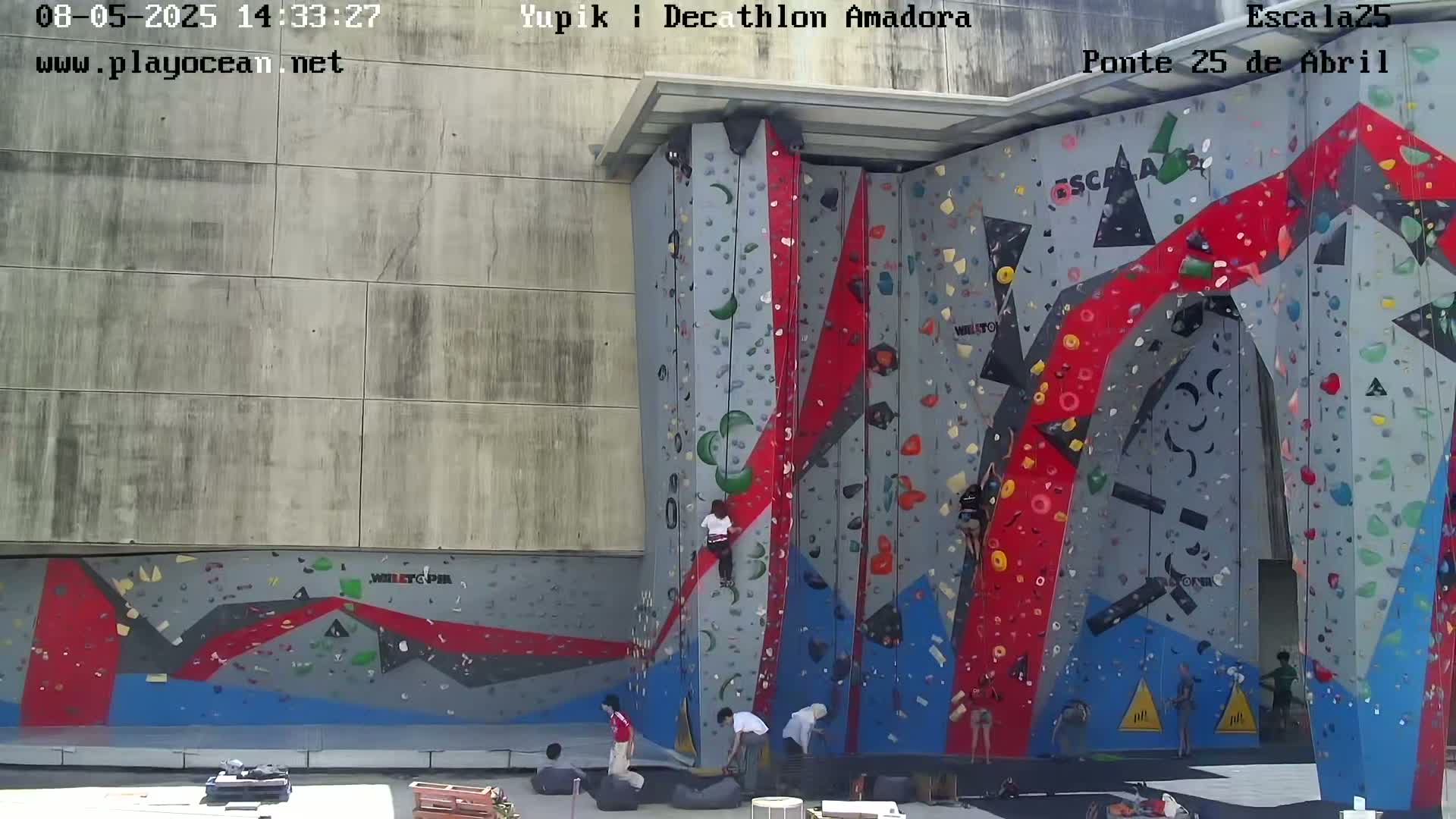 Several people climb a large, colorful indoor climbing wall on a sunny day.