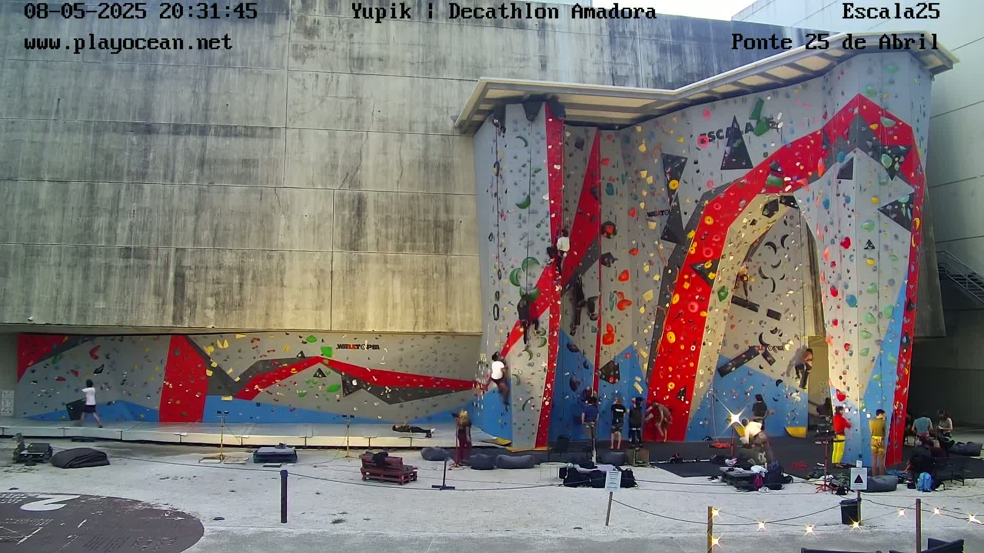 Several people are climbing and watching others climb a large, colorful outdoor climbing wall at night.