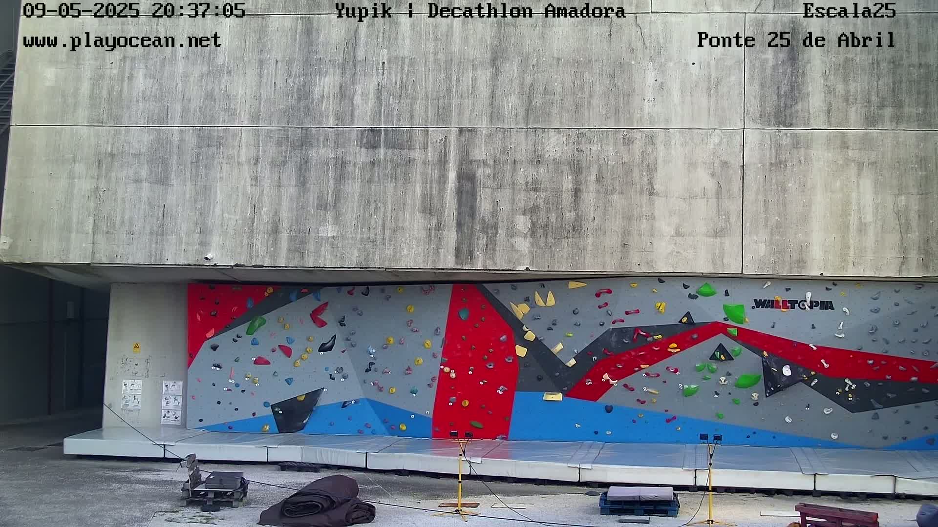 A large outdoor climbing wall with various colored holds is situated beneath a concrete overhang.