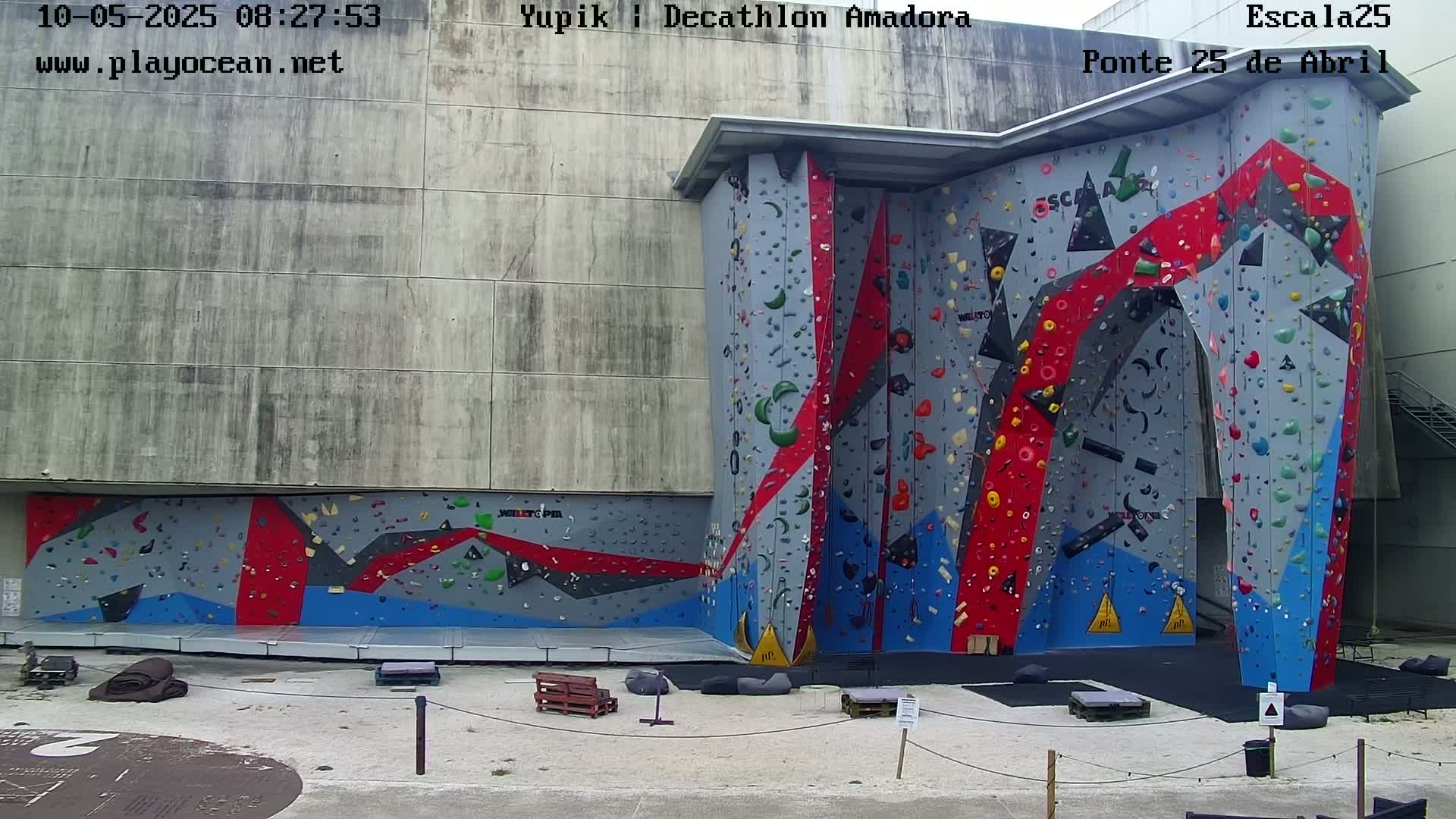 An outdoor climbing wall with various colored holds and a surrounding paved area.