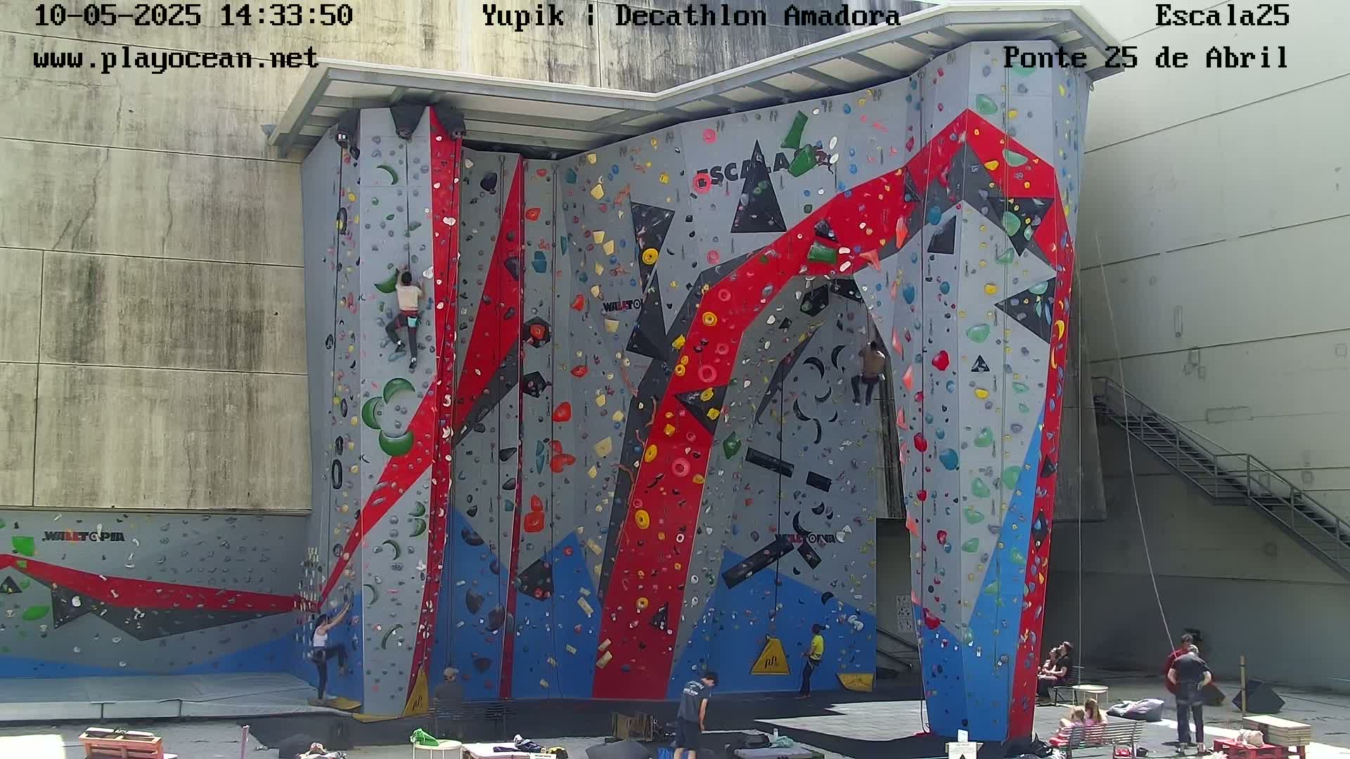 A large, colorful indoor climbing wall stands against a light-grey building under a sunny sky, with several people climbing and watching.