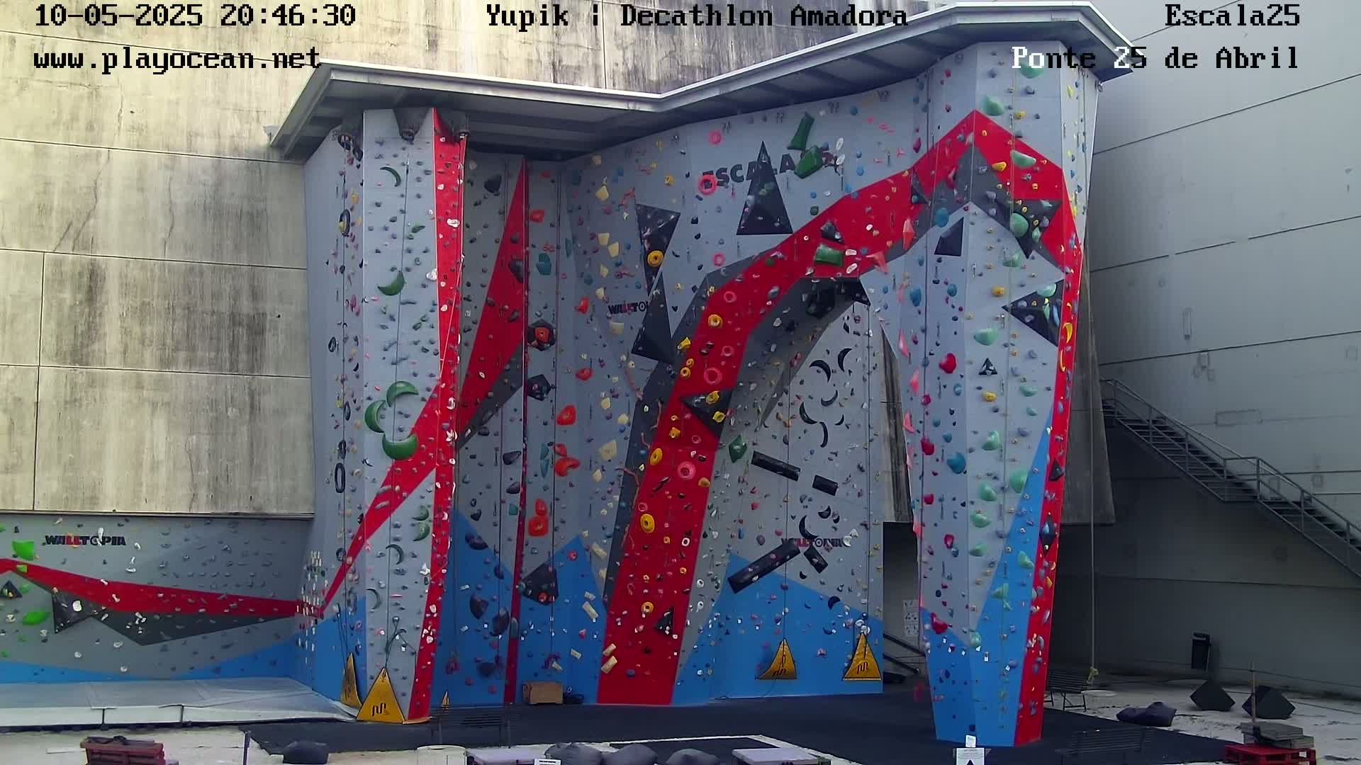 A large indoor climbing wall with various colored holds and a gray background is visible under a daytime sky.