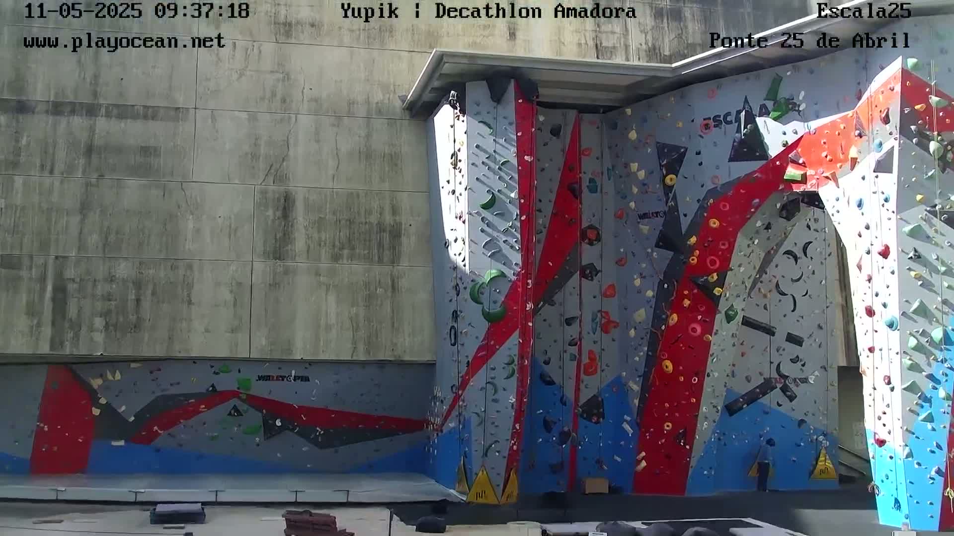 A large, colorful indoor climbing wall is situated against a concrete building in sunny conditions.