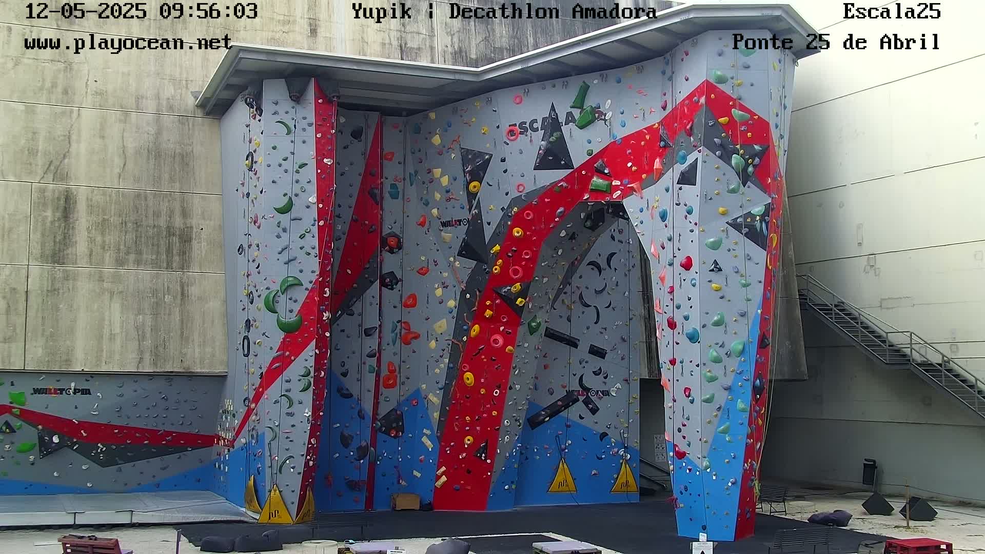 A large, colorful indoor climbing wall with various holds is situated against a light gray building.