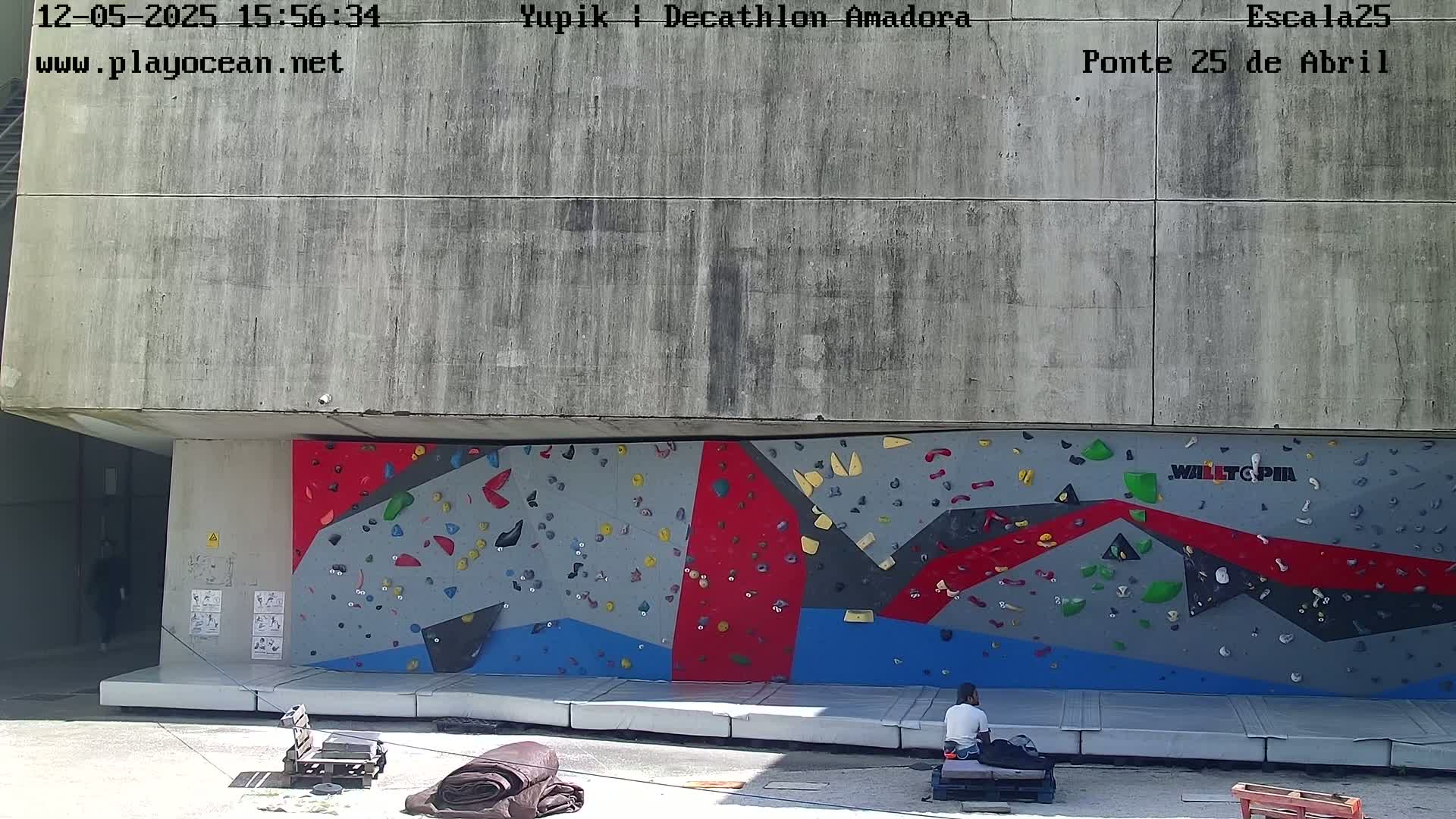 A person sits near an outdoor climbing wall on a sunny day.