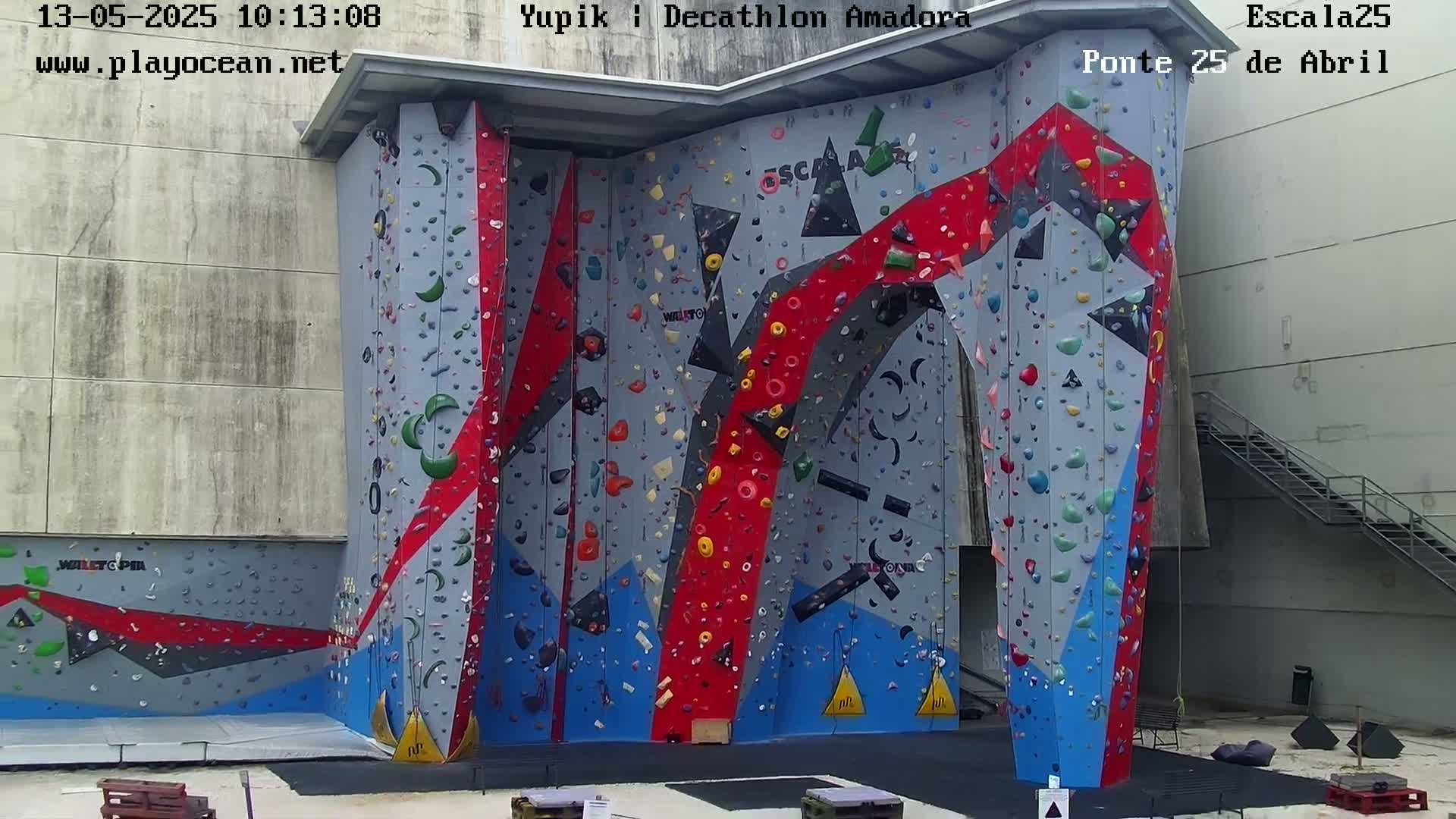A large, colorful indoor climbing wall with various holds stands against a light-grey building.