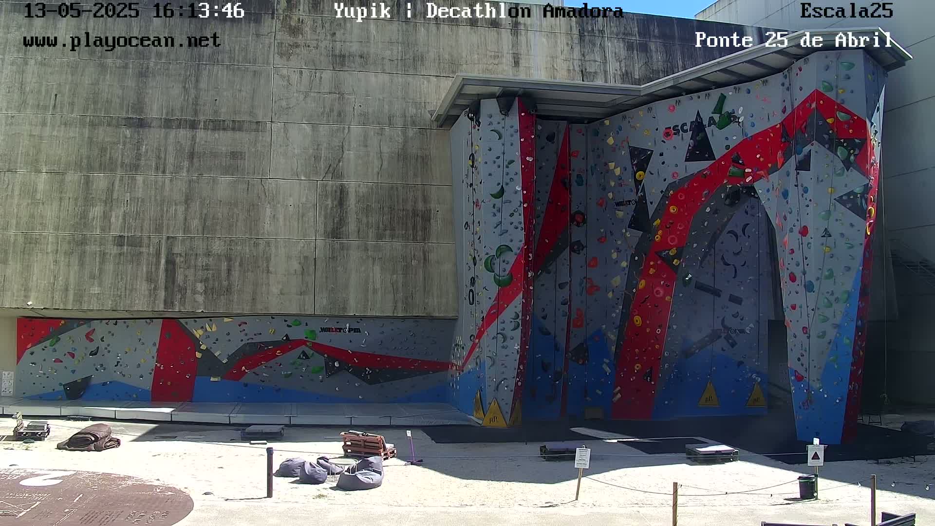 A large outdoor climbing wall with red, gray, and blue sections is situated against a concrete building on a sunny day.