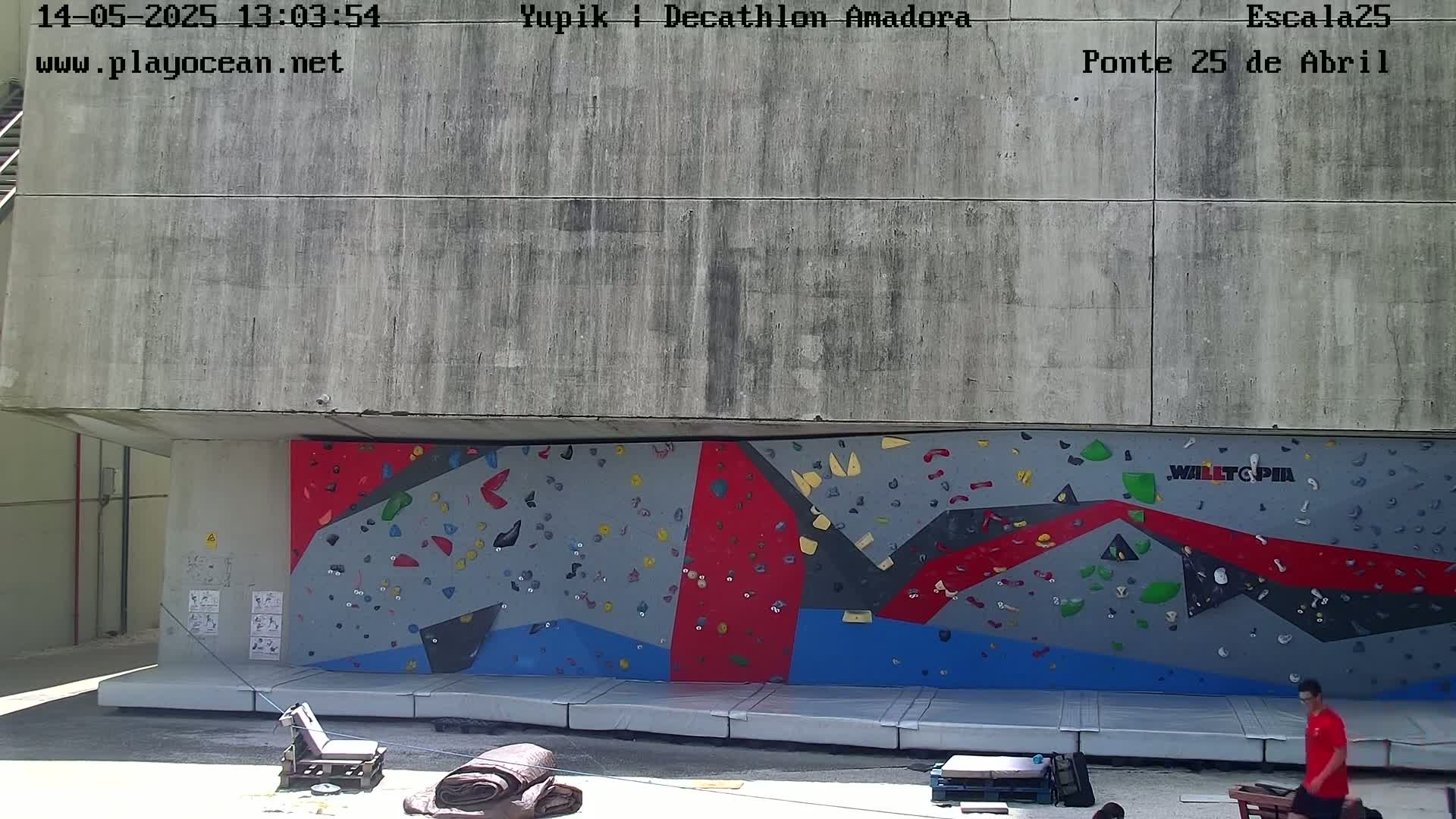 A person stands near a large, colorful climbing wall outdoors on a sunny day.