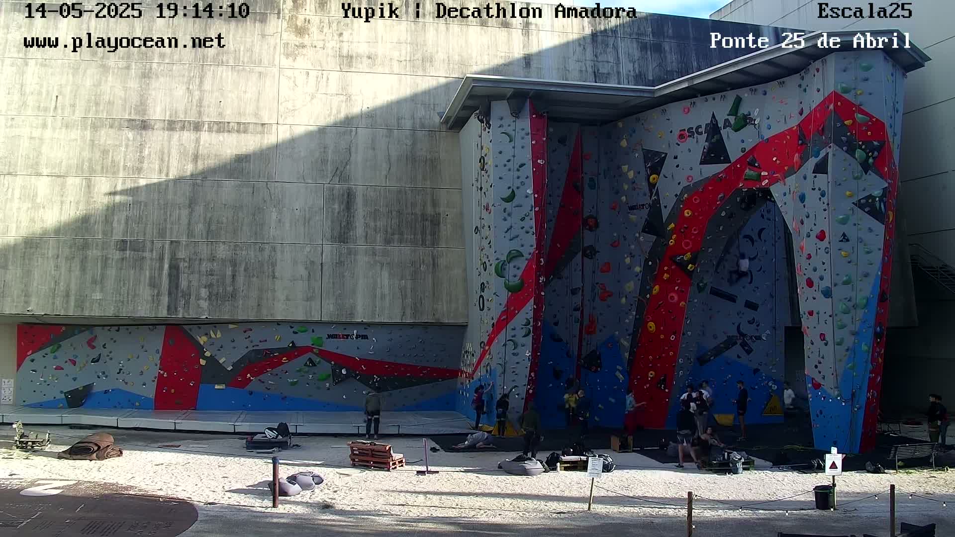A large outdoor climbing wall with various colored holds is situated next to a concrete building, under sunny conditions with a few people climbing and resting nearby.