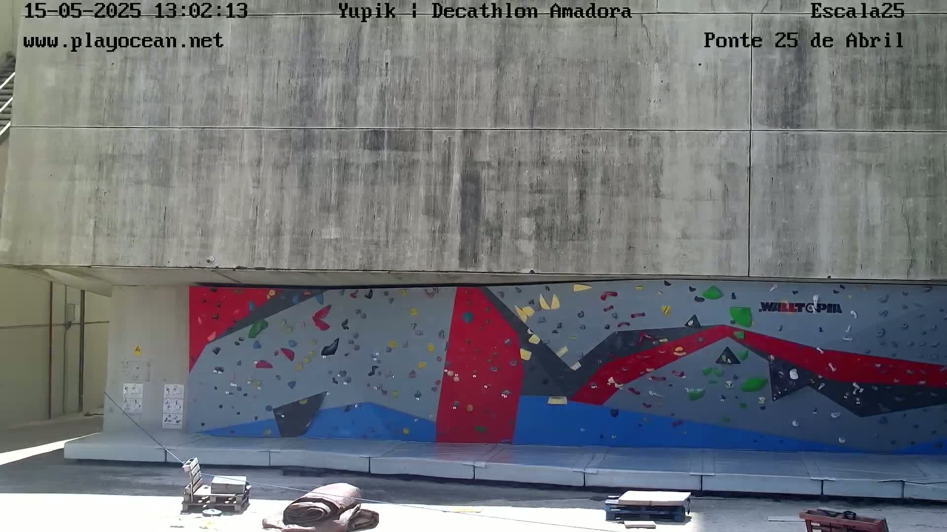 An outdoor climbing wall with red, gray, and blue sections is situated beneath a concrete overhang on a sunny day.