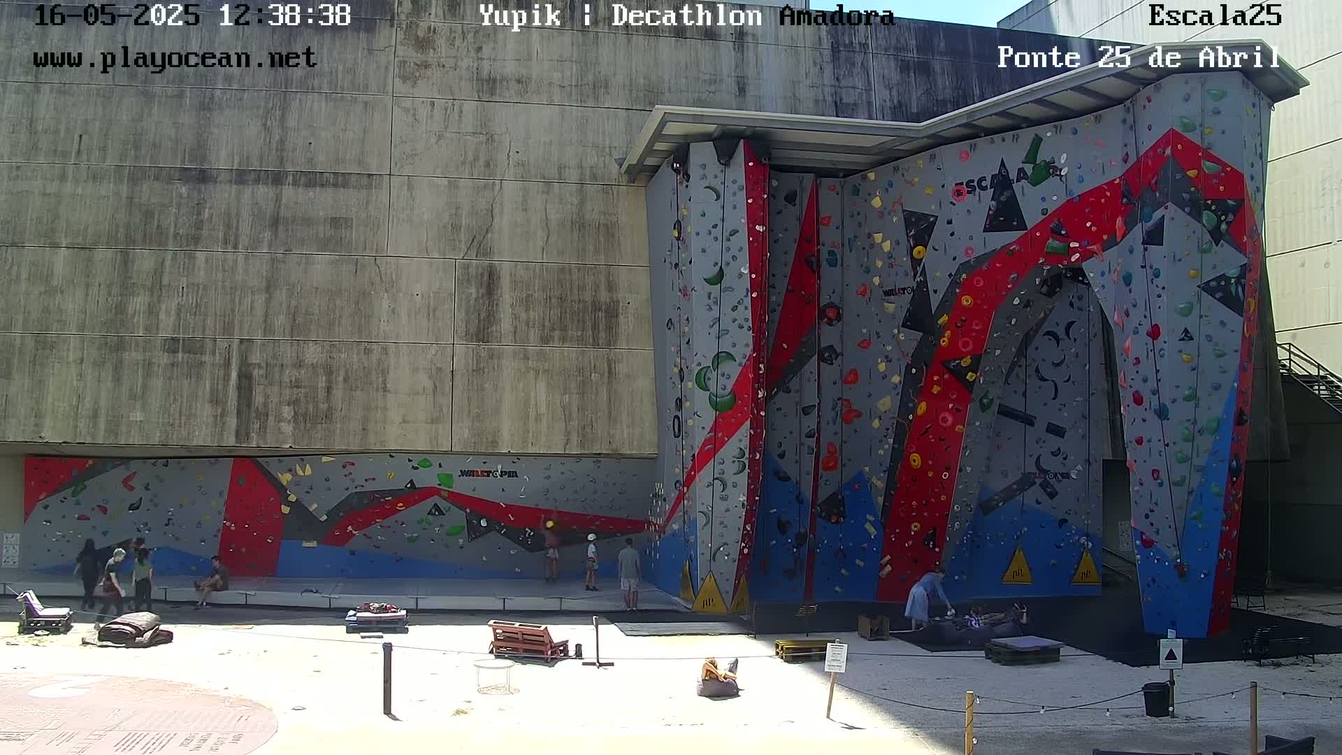 A large, colorful outdoor climbing wall is surrounded by a few people on a sunny day.