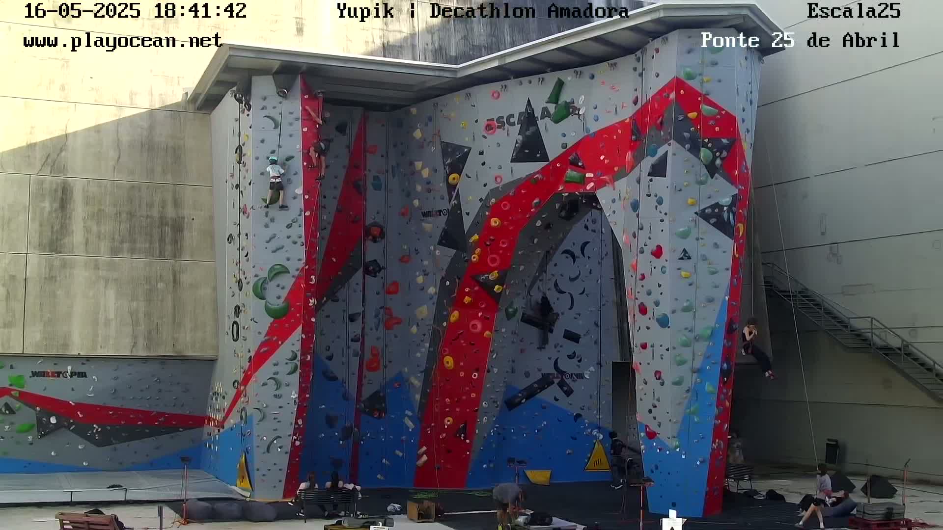 Several people are rock climbing on a large, colorful outdoor climbing wall.