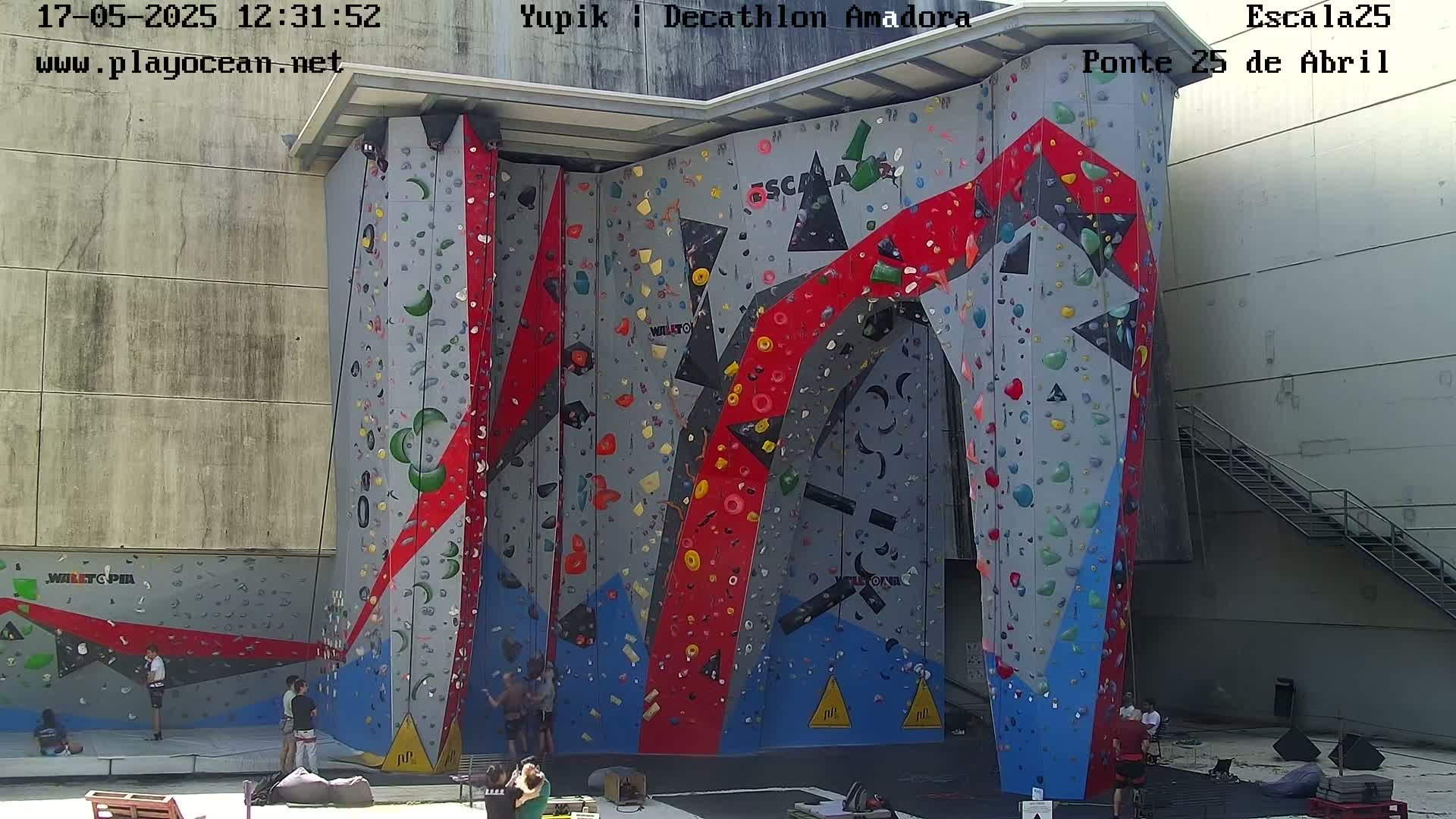 A large, colorful outdoor climbing wall with several people at its base on a sunny day.