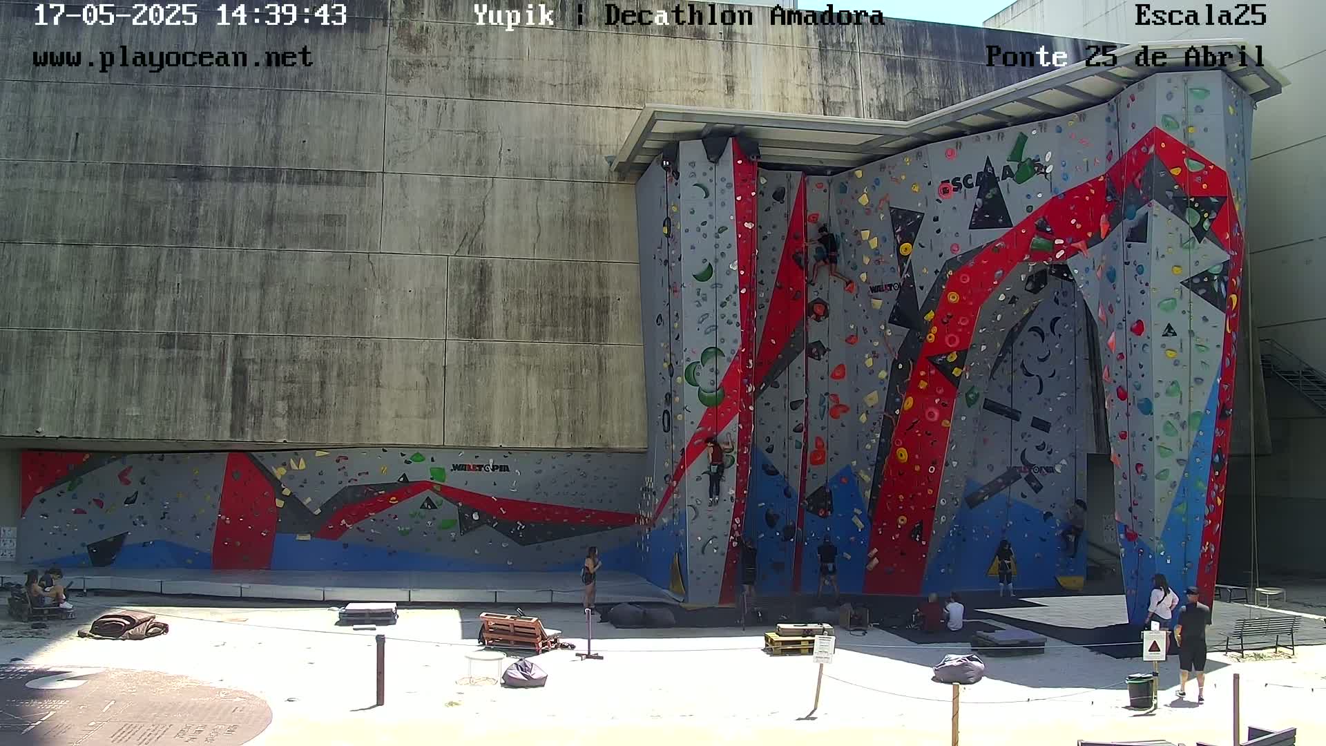 A large, colorful outdoor climbing wall is situated against a concrete building on a sunny day, with several people climbing and resting nearby.