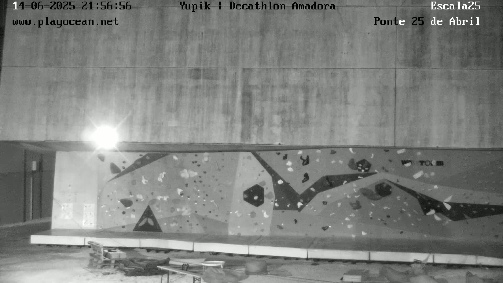 A large, colorful outdoor climbing wall is situated against a concrete building on a sunny day, with several people climbing and resting nearby.